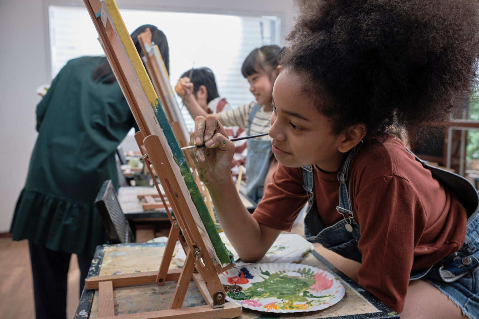 Young girl concentrates on acrylic color picture painting on canvas with students group in art classroom